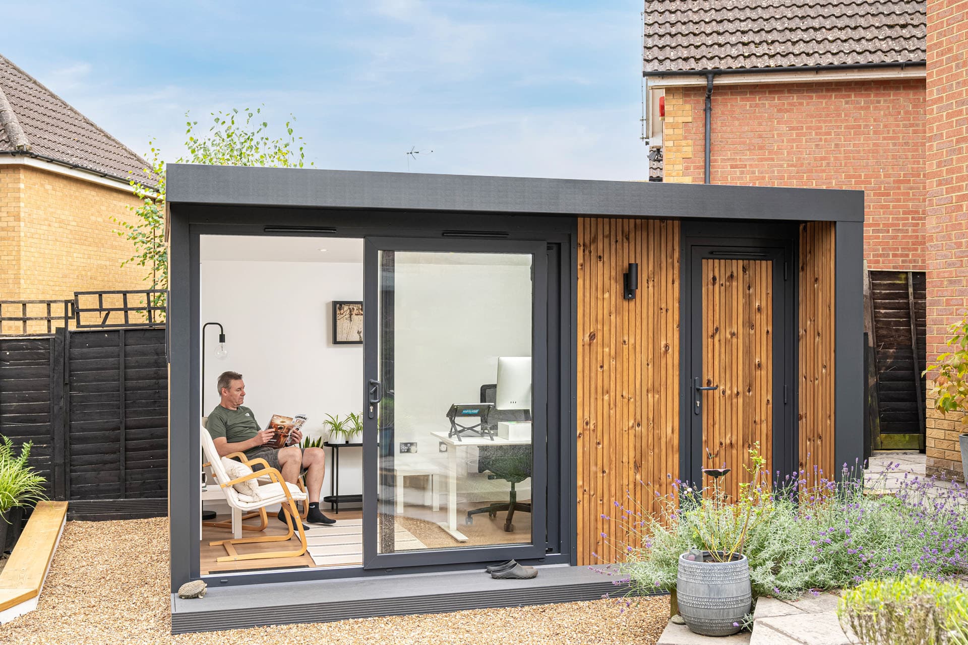 A contemporary backyard garden room featuring hardwood paneling and a sleek black frame. Large sliding glass doors in the garden area offer a comfortable interior arrangement with plants, a chair, and a small table. The outdoor space includes a patio that is kept up, a pathway made of gravel, and a dining table and chairs that are covered by an umbrella. The area is bordered by a lavender bush and lush vegetation, which add to the cozy and natural ambiance.